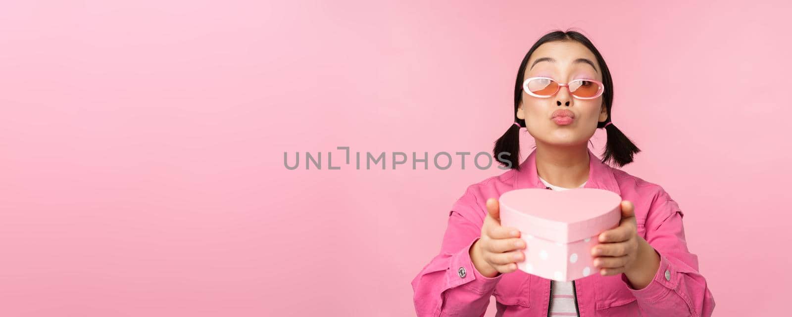 Cute asian girl giving you gift in heart shaped box, kissing and smiling, concept of holiday and celebration, standing over pink background.