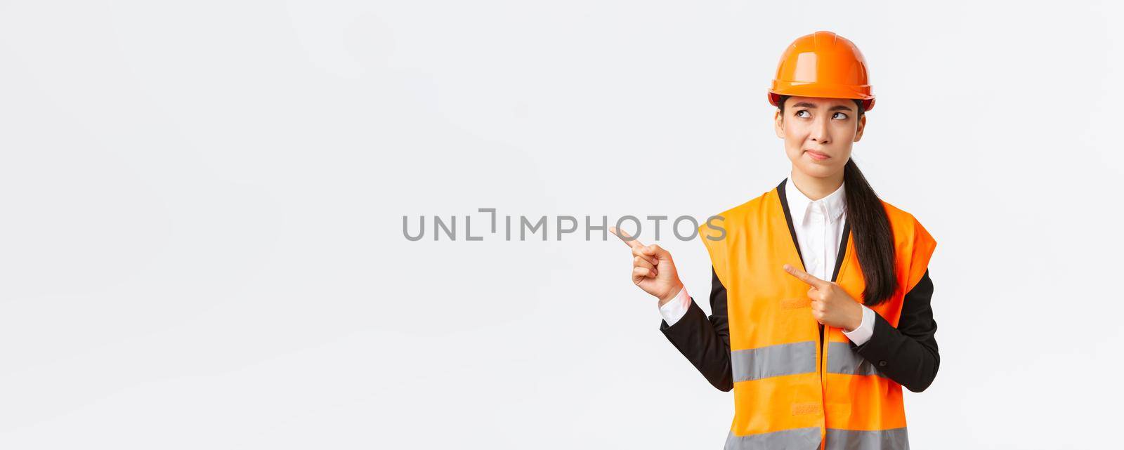 Building, construction and industrial concept. Indecisive asian female architect being unsure, wearing reflective clothing and helmer, smirk as pointing and looking upper left corner puzzled by Benzoix