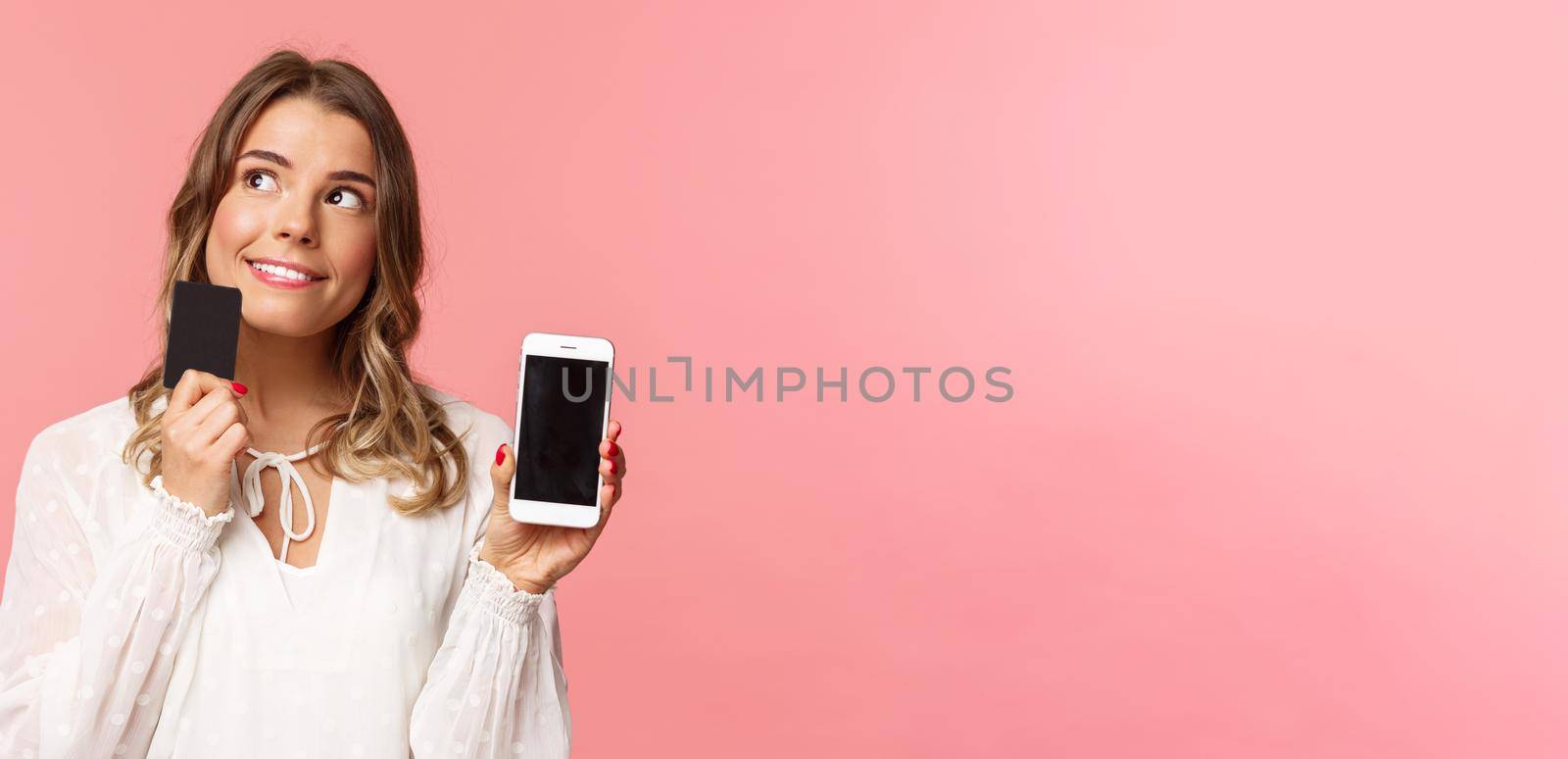 Finance, shopping and technology concept. Close-up portrait of thoughtful, dreamy attractive blond girl in white dress, thinking what buy, order something online shop, hold smartphone and credit card by Benzoix