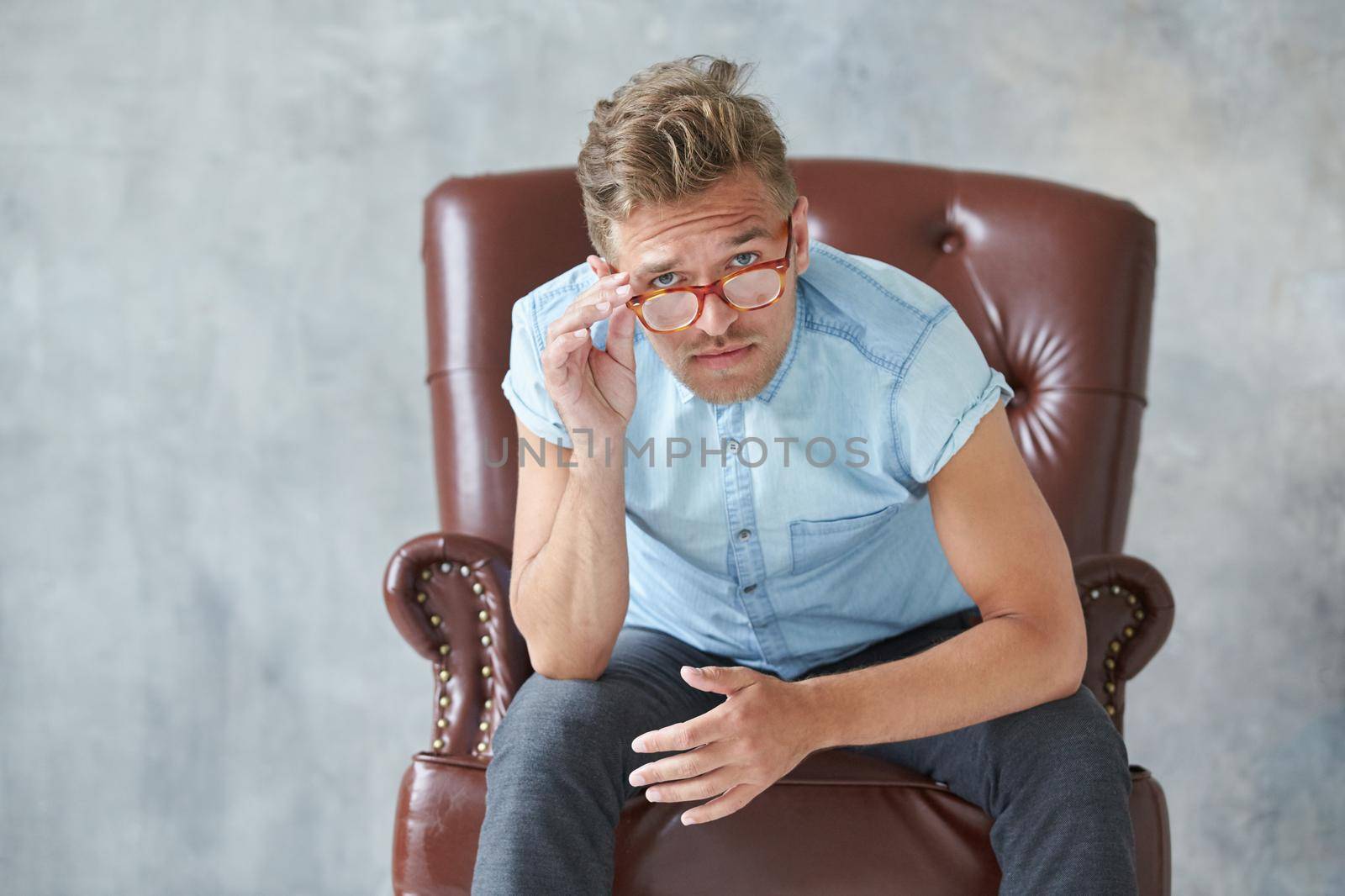 Portrait of a stylish intelligent man stares into the camera, small unshaven, charismatic, blue shirt, sitting on a brown leather chair, dialog, negotiation, short sleeve, brutal, hairstyle. High quality photo