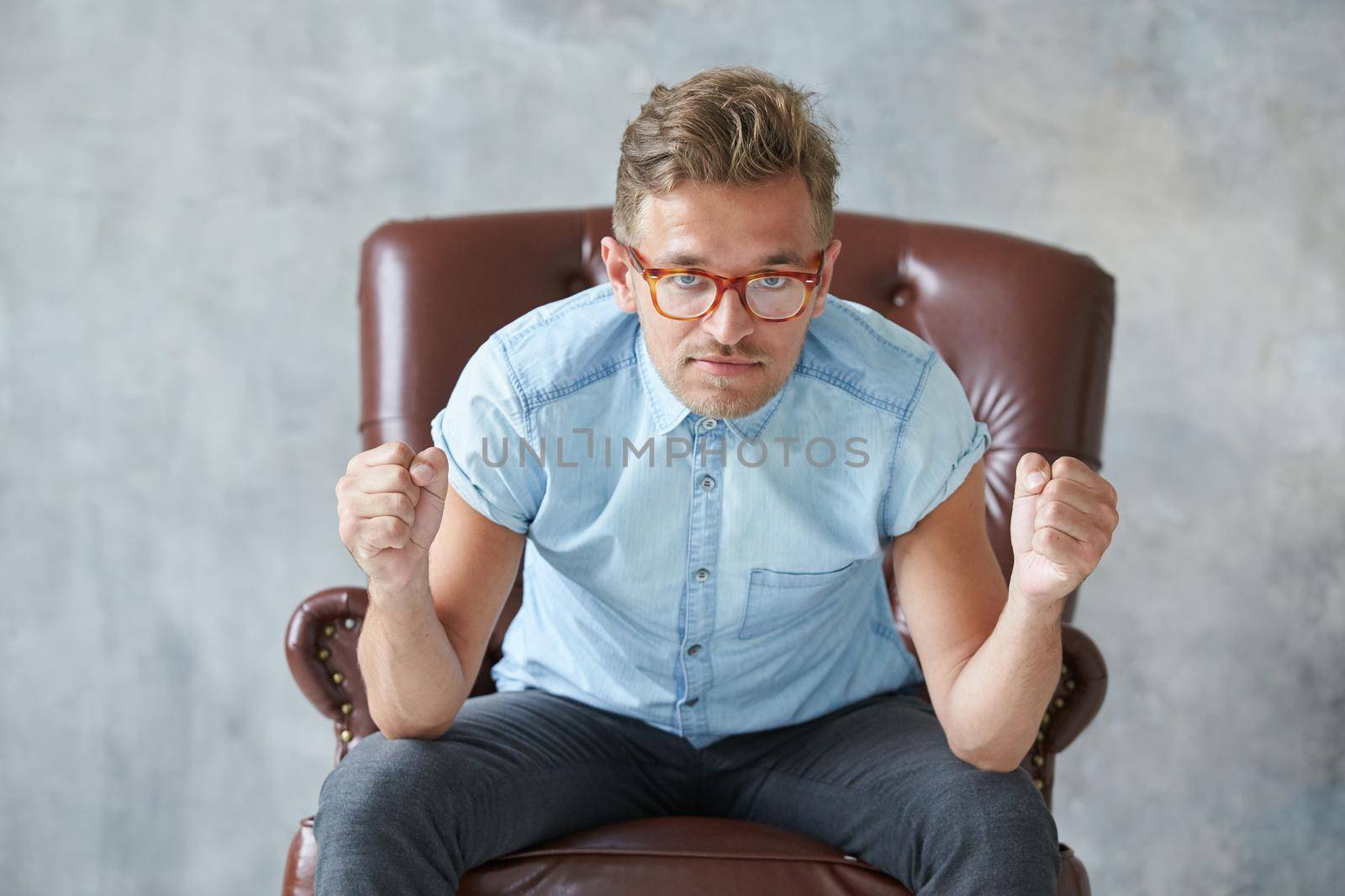 Portrait of a stylish intelligent man stares into the camera, small unshaven, charismatic, blue shirt, sitting on a brown leather chair, dialog, negotiation, short sleeve, brutal, hairstyle. High quality photo