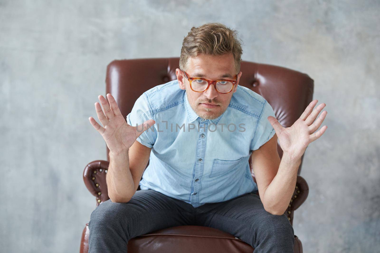 Portrait of a stylish intelligent man stares into the camera, small unshaven, charismatic, blue shirt, sitting on a brown leather chair, dialog, negotiation, short sleeve, brutal, hairstyle. High quality photo