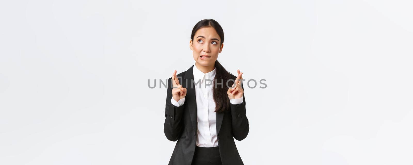 Hopeful asian businesswoman looking worried, biting lip and cross fingers good luck, making wish, anticipating big contract or deal, worried about outcome, standing white background.