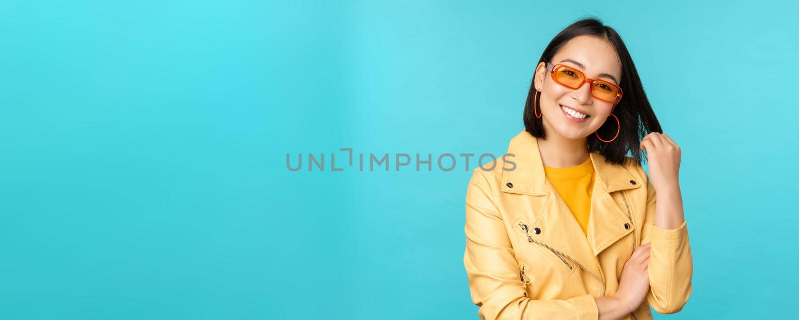 Young stylish asian woman in sunglasses smiling, playing with her haircut and looking happy, posing against blue background by Benzoix