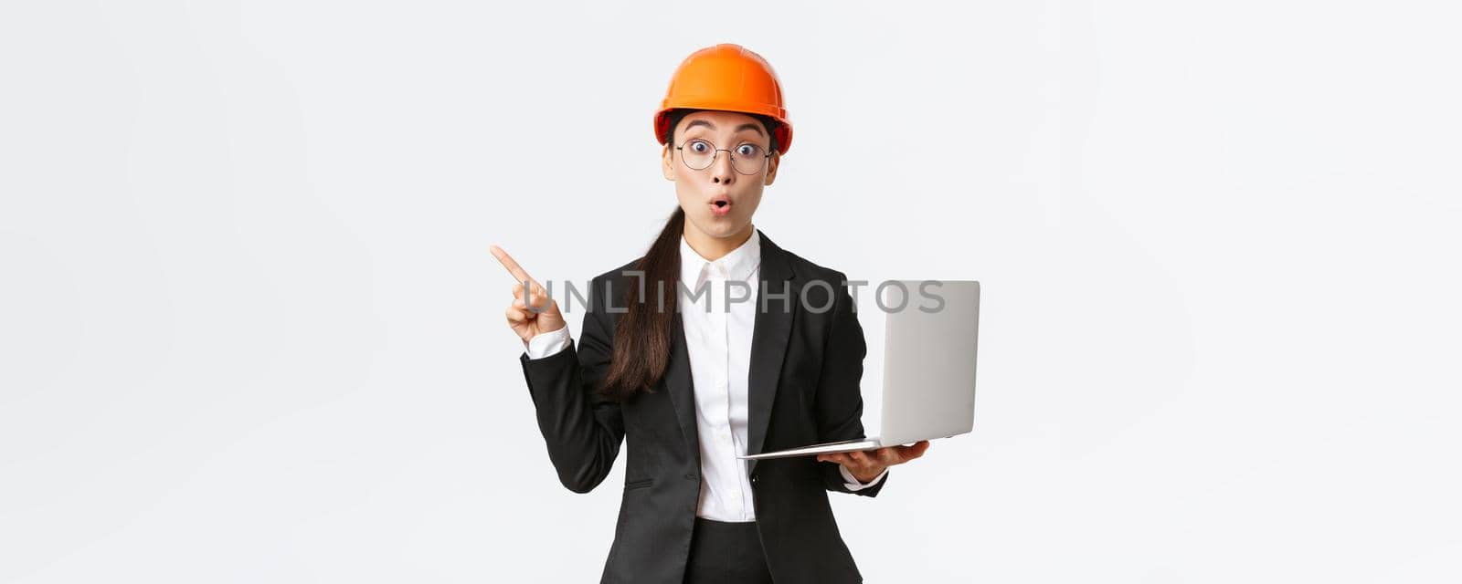 Impressed asian female entrepreneur at factory wearing safety helmet and business suit, pointing finger left and holding laptop computer, showing diagram at enterprise, white background.