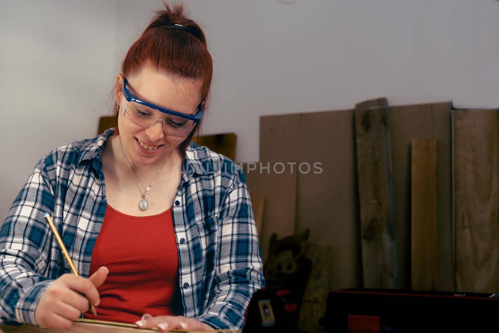 Young woman carpenter smilling, with red hair, working on wood design in a small carpentry workshop, dressed in blue checked shirt and red t-shirt. Young businesswoman handcrafting a piece of wood in her small business. Warm light indoors, background with wooden slats. Horizntal.