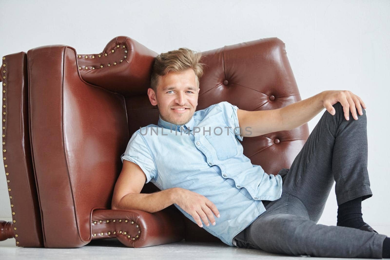 Brutal man lying next to a brown leather armchair , armchair lying on its side on the floor , a man holds hands behind the chair. High quality photo