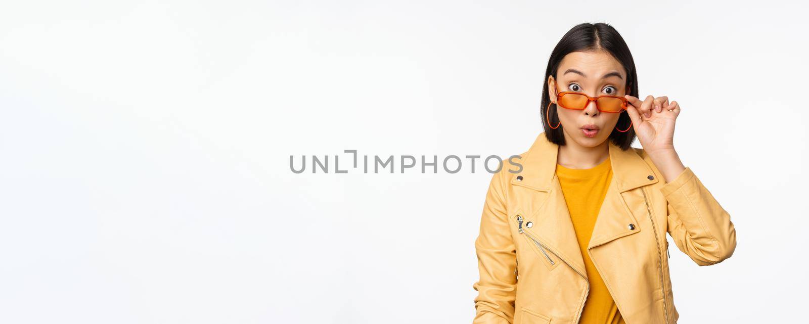 Portrait of asian brunette woman in stylish sunglasses, looks surprised and impressed at camera, checking out big news, wow face expression, standing over white background.