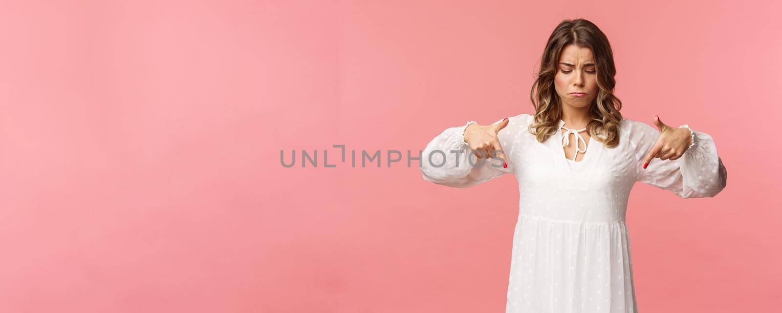 Gloomy, jealous and upset cute blond european woman in white trendy spring dress, sulking as pointing and looking down, see something expensive, cant efford it, strand pink background.