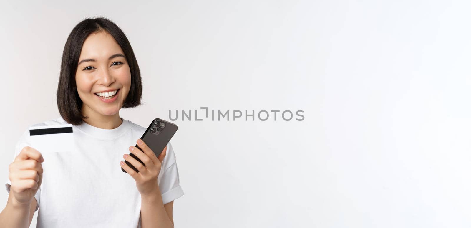 Smiling asian girl using mobile phone, showing credit card, concept of contactless payment, online shopping, standing over white background.