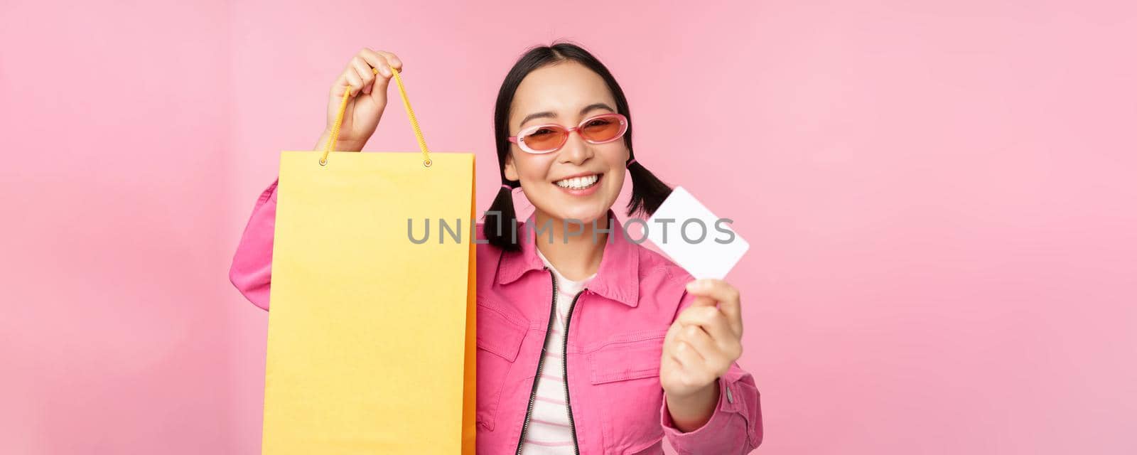 Happy young asian woman showing credit card for shopping, holding bag, buying on sale, going to the shop, store, standing over pink background.