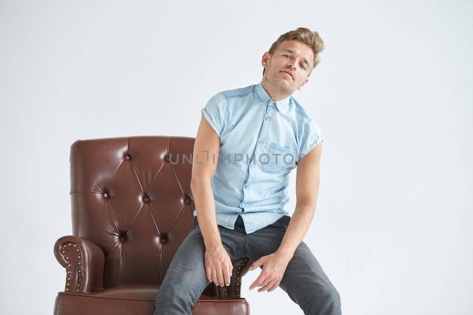 Portrait of a stylish intelligent man, small unshaven, charismatic, blue shirt, sitting on a brown leather chair, dialog, negotiation, short sleeve, brutal, hairstyle, emotions, indifference, fatigue. High quality photo