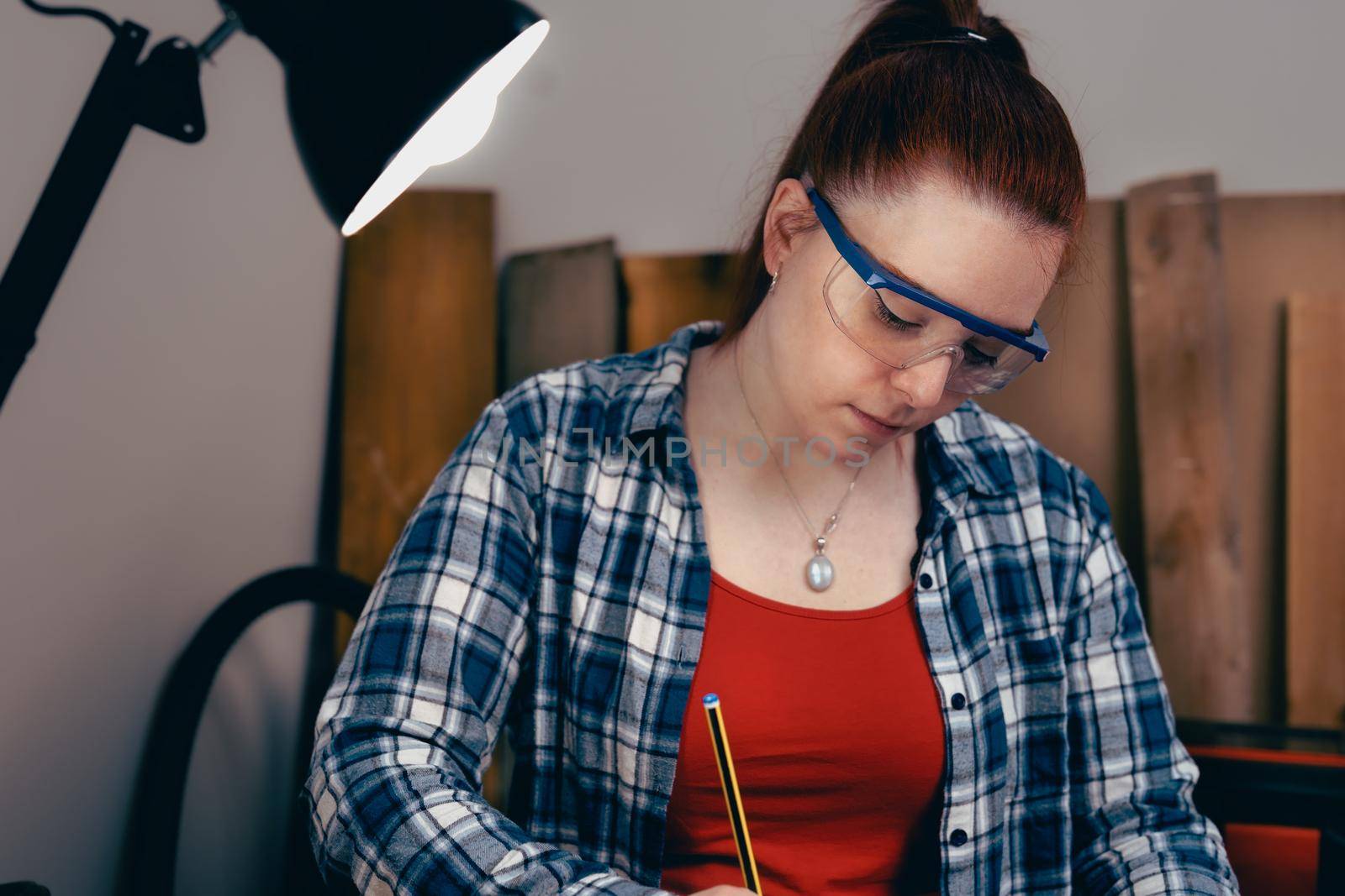 Young carpenter with red hair, working on wood design in a small carpentry workshop, dressed in blue checked shirt and red t-shirt. Young businesswoman handcrafting a piece of wood in her small business. Warm light indoors, background with wooden slats. Horizntal.