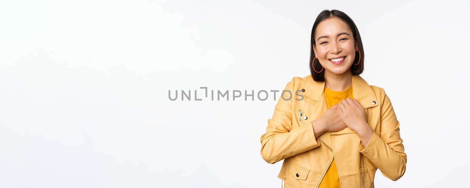 Portrait of smiling, beautiful asian girl looking with appreciation, thank you gesture, holding hands on heart flattered, standing over white background.