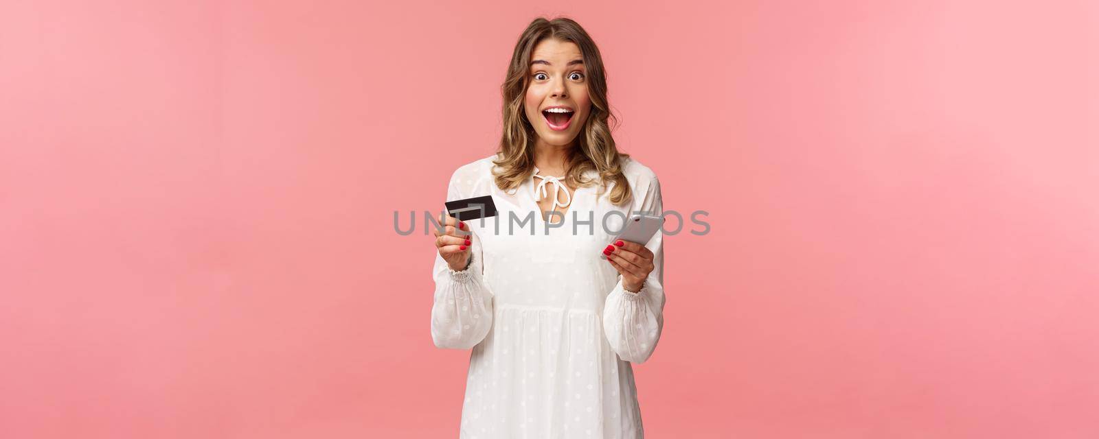 Portrait of excited and amused, happy smiling caucasian woman making order internet, shopping online with mobile phone, enter credit card, billing info, look camera amazed, pink background.