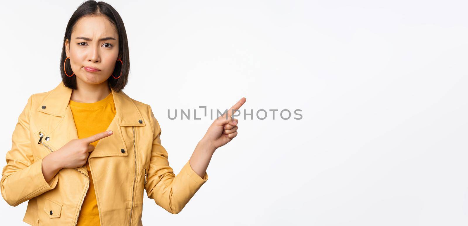 Disappointed asian girl frowning, sulking upset, pointing fingers right at advertisement, standing over white background by Benzoix