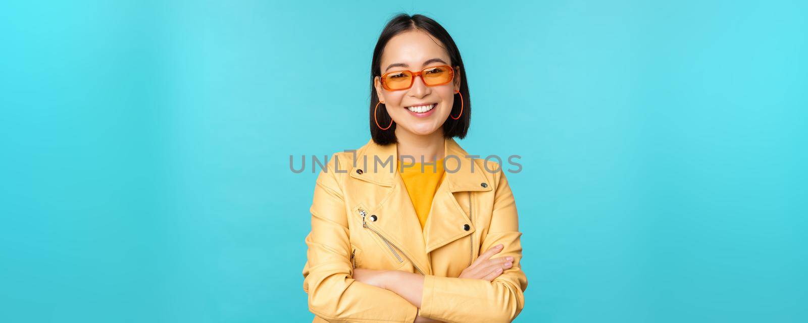 Stylish korean woman in sunglasses and yellow jacket, cross arms, smiling and looking confident at camera, standing against blue background.