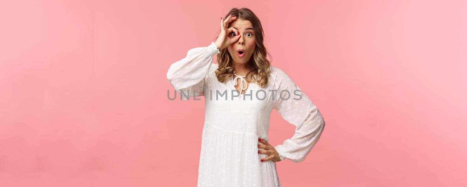 Portrait of amazed and intrigued young blond girl seeing something awesome, look from okay sign with startled excited expression, fold lips say wow, pop eyes at camera, pink background.