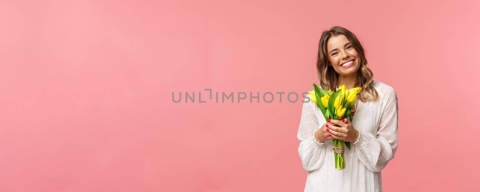 Holidays, beauty and spring concept. Portrait of lovely caucasian blond girl in white dress, smiling upbeat, holding yellow tulips, having perfect romantic date with flowers as gift, pink background by Benzoix