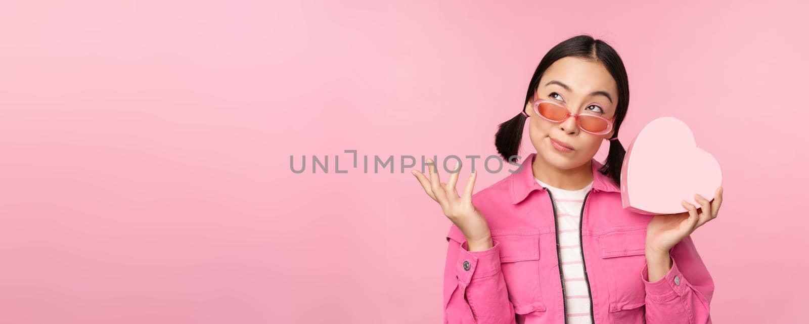 Image of stylish asian girlfriend in sunglasses, guessing whats inside gift box, heart-shaped present, standing over pink background.