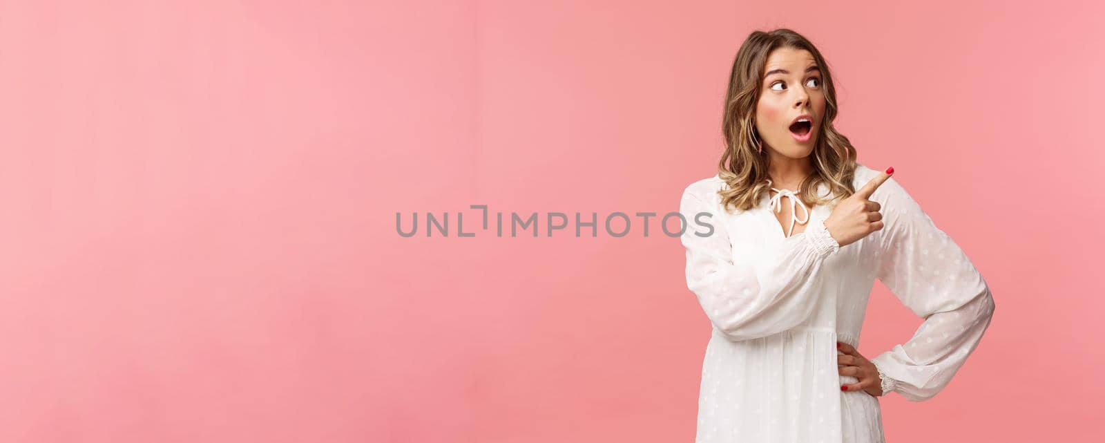 Surprised and excited good-looking blond girl in white cute dress, open mouth in amazement, look curious pointing upper right corner astonished, see something awesome, pink background.