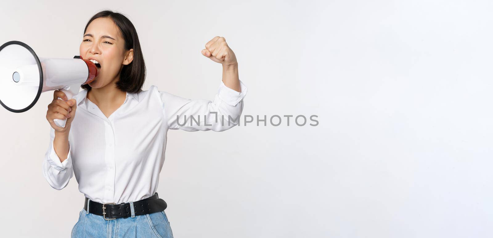 Enthusiastic asian woman, girl activist shouting at protest, using megaphone, looking confident, talking in loudspeaker, protesting, standing over white background.