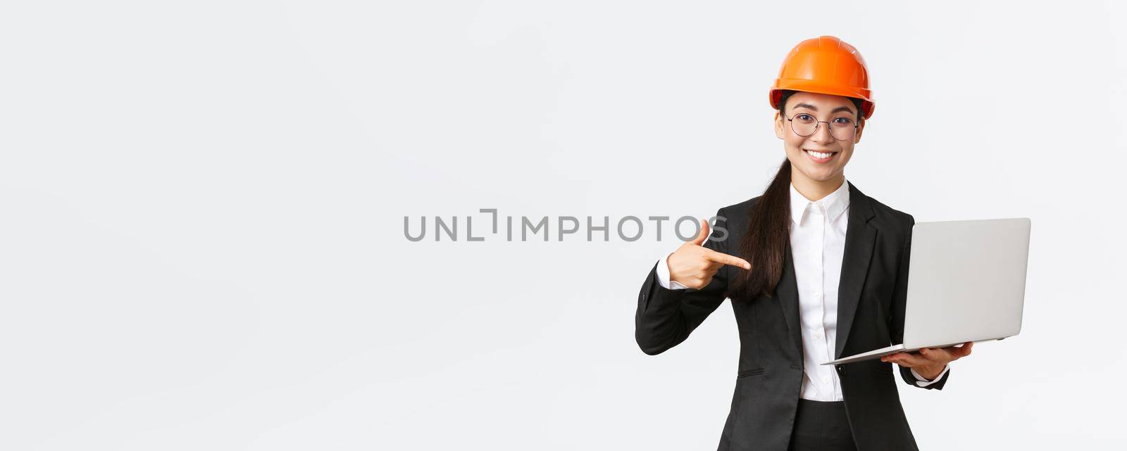 Smiling pleased asian female chief engineer showing chart with enterprise profit, making presentation during investors meeting, wearing safety helmet, pointing at laptop screen, white background.