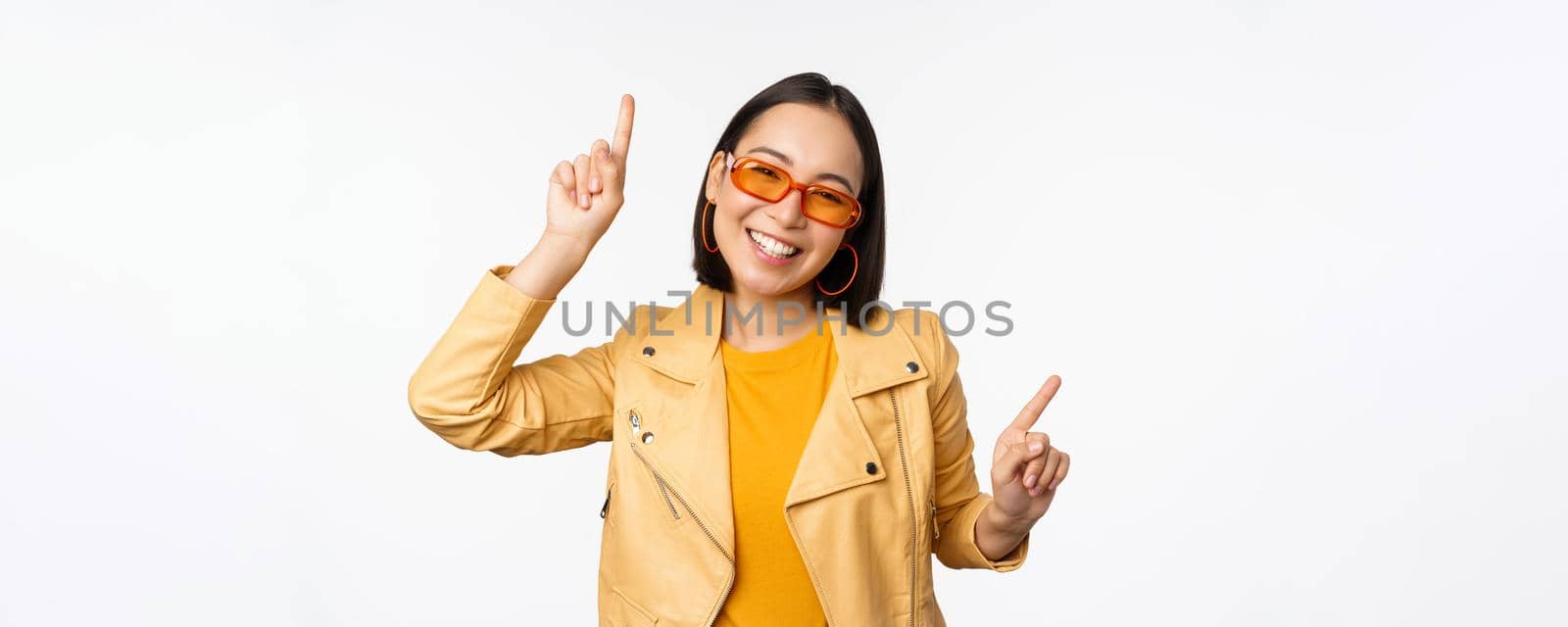 Smiling asian brunette woman in sunglasses, pointing fingers sideways, left and right, showing variants, laughing and dancing, wearing sunglasses, white background.