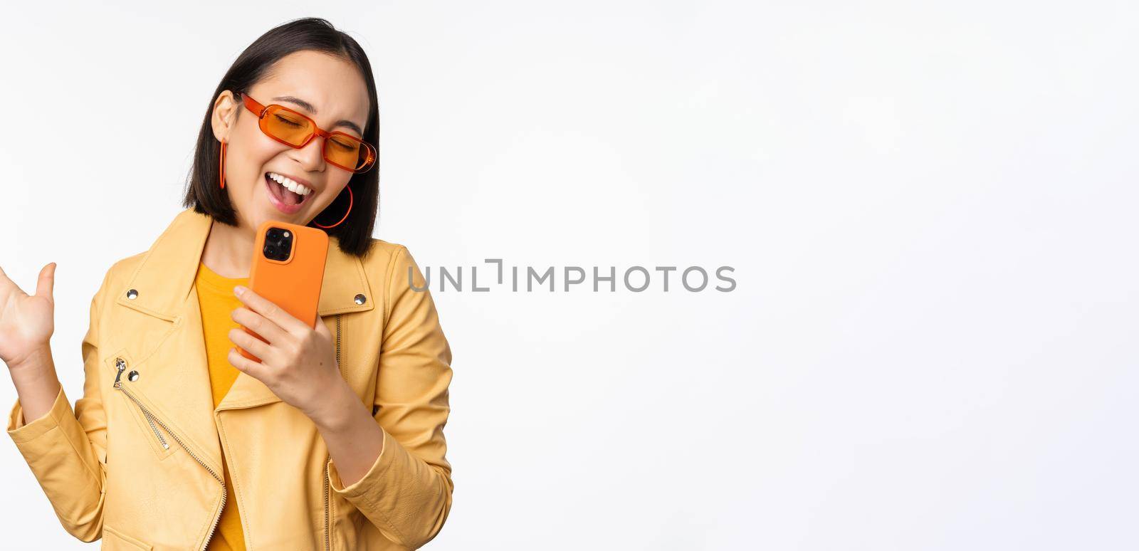 Technology and people concept. Happy dancing asian girl in sunglasses, using smartphone, singing and smiling, standing over white background.