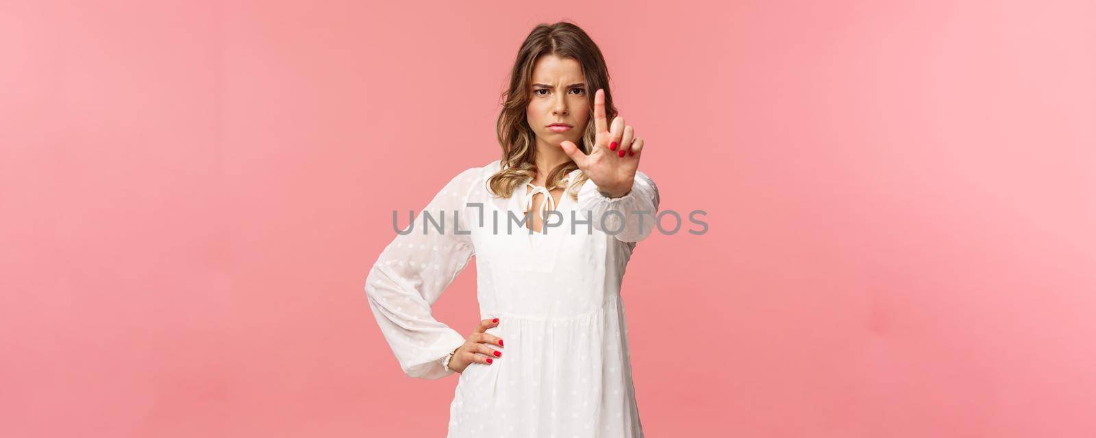 Stop, enough. Portrait of serious-looking assertive and strong blond girl in white dress, extend hand towards camera with one finger raised in warning, frowning angry, forbid and prohibit action.
