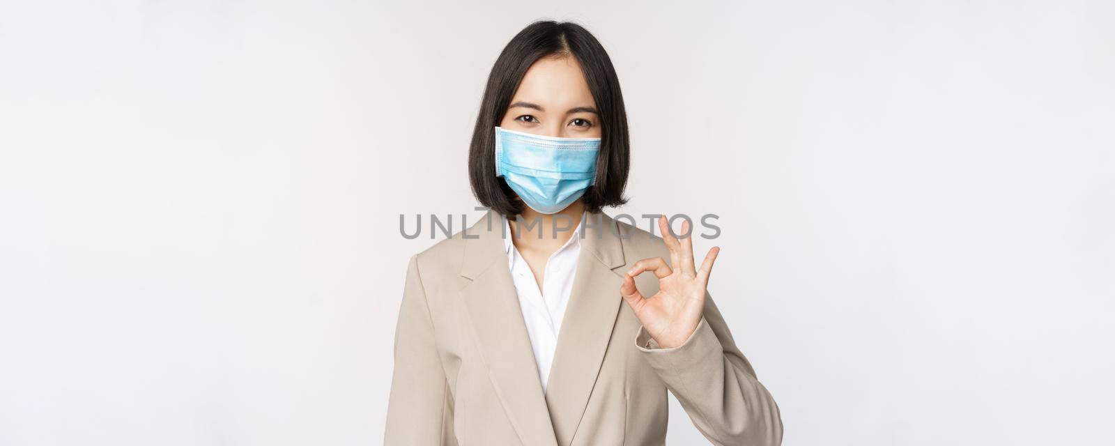 Coronavirus and workplace concept. Image of asian saleswoman, company worker in medical mask showing okay sign, smiling pleased, white background by Benzoix