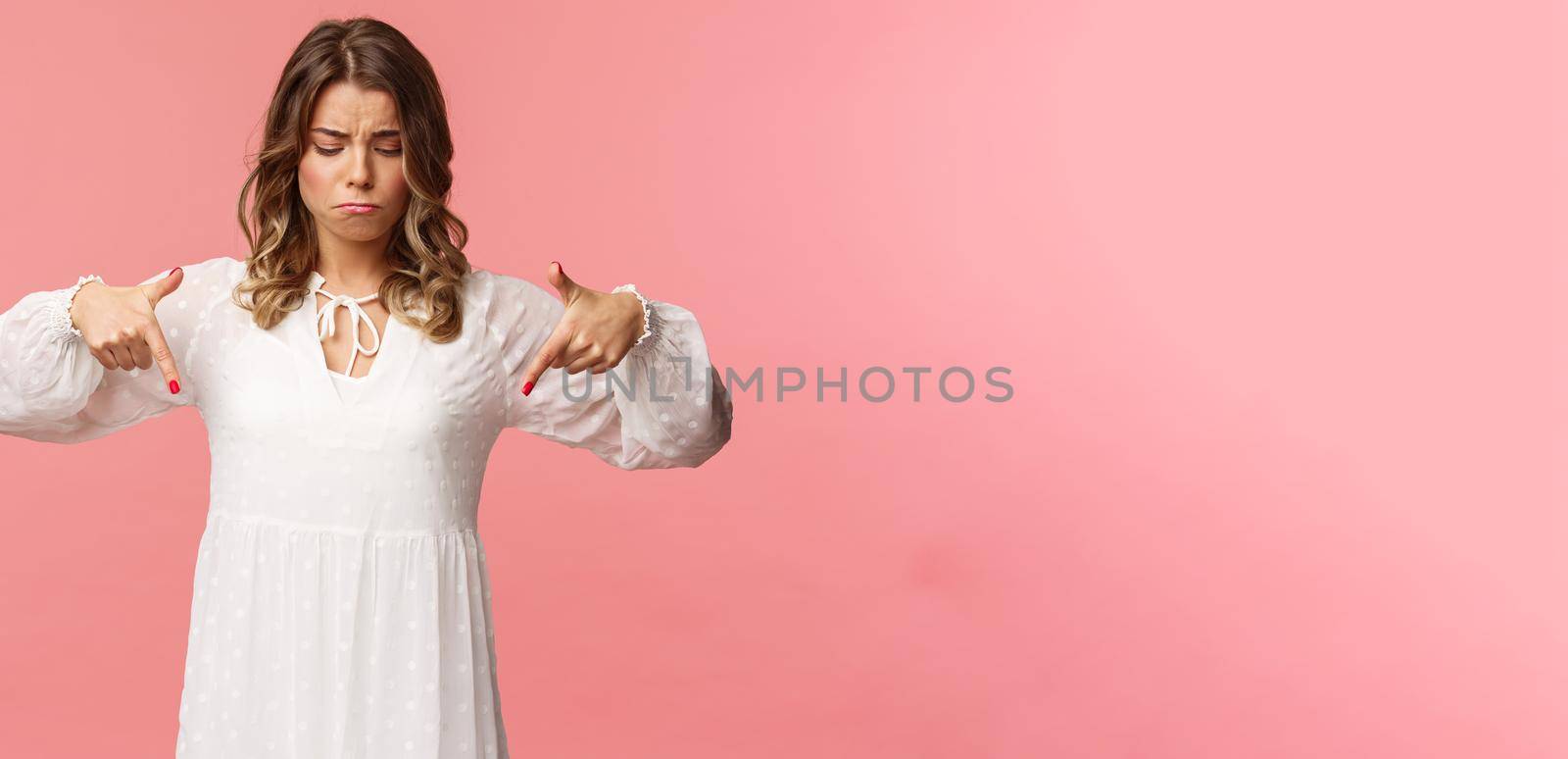 Gloomy, jealous and upset cute blond european woman in white trendy spring dress, sulking as pointing and looking down, see something expensive, cant efford it, strand pink background.