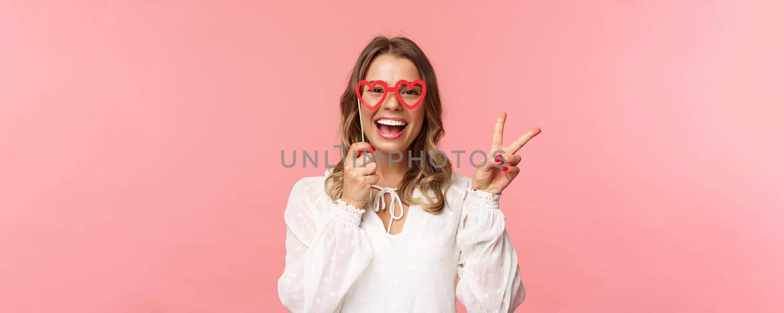 Spring, happiness and celebration concept. Close-up portrait of upbeat blond girl at party, wear white dress holding heart-shaped glasses mask over eyes and make peace sign cheerful by Benzoix