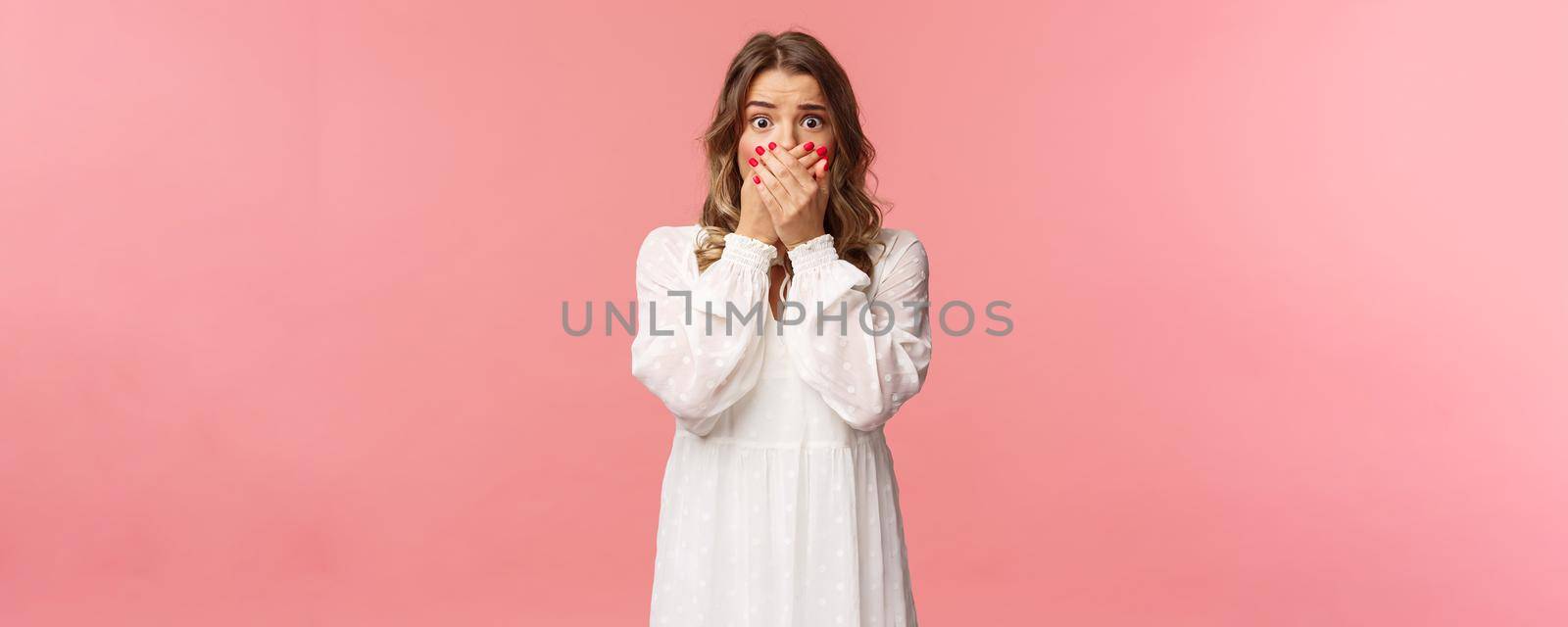 Shocked and anxious young concerned pretty girl in white dress, gasping close mouth with hands, look worried and startled, express fear or empathy with eyes, standing pink background.