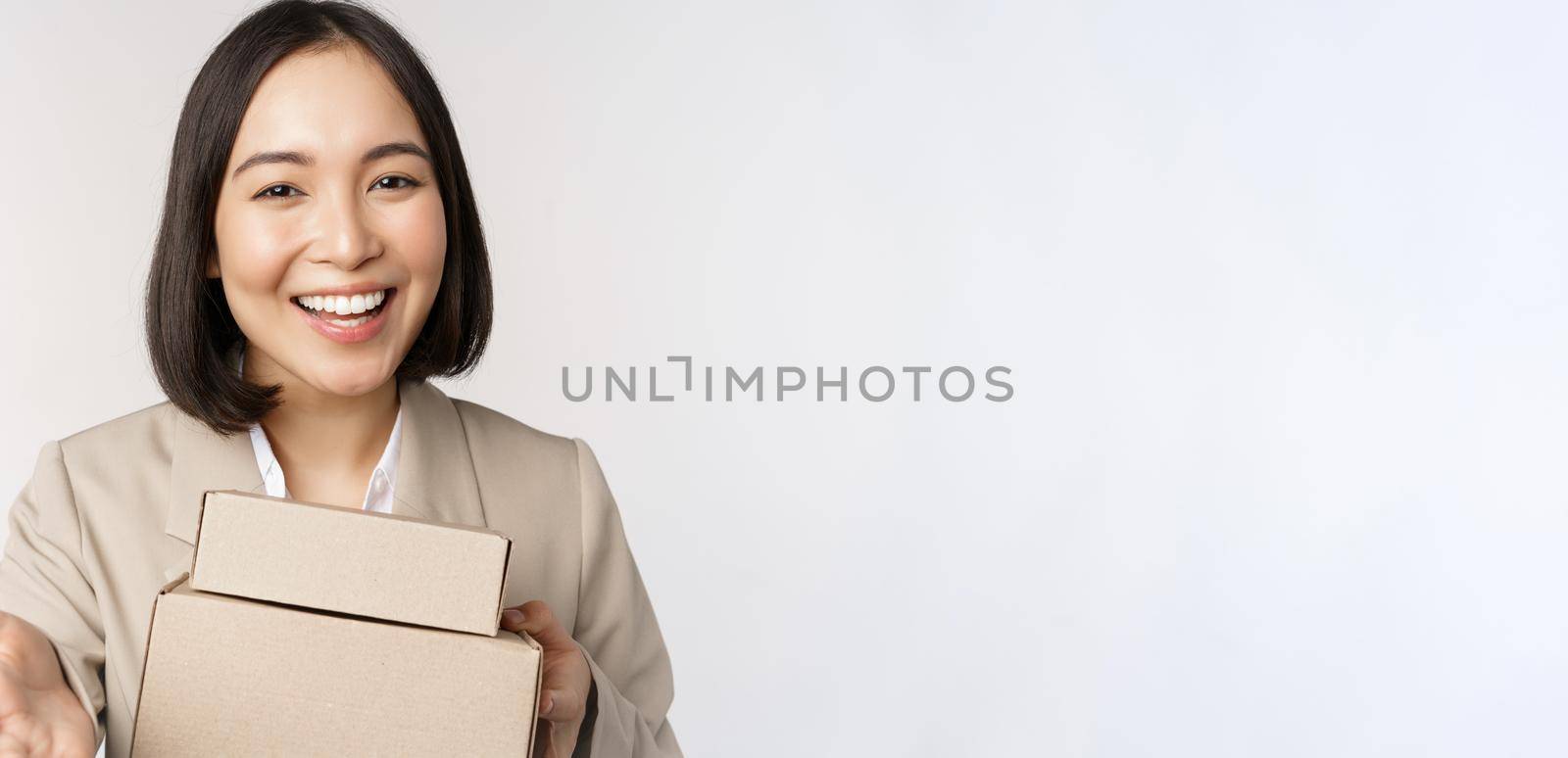 Portrait of business woman, asian saleswoman pointing at you, giving boxes with orders, standing in suit over white background.