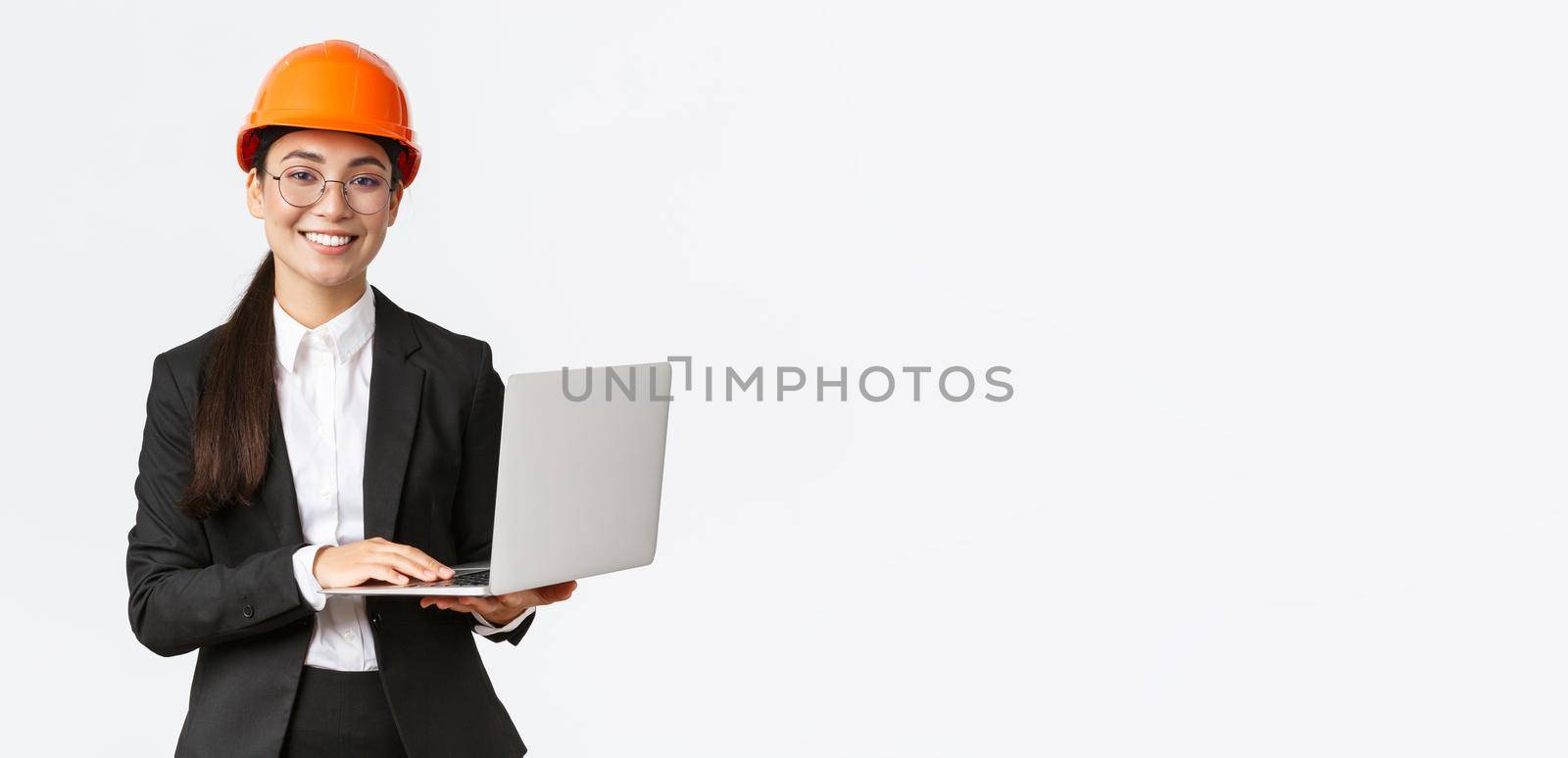 Smiling professional female saleswoman showing construction works on new house. Asian industrial engineer in safety helmet and business suit using laptop computer, enter data from factory.