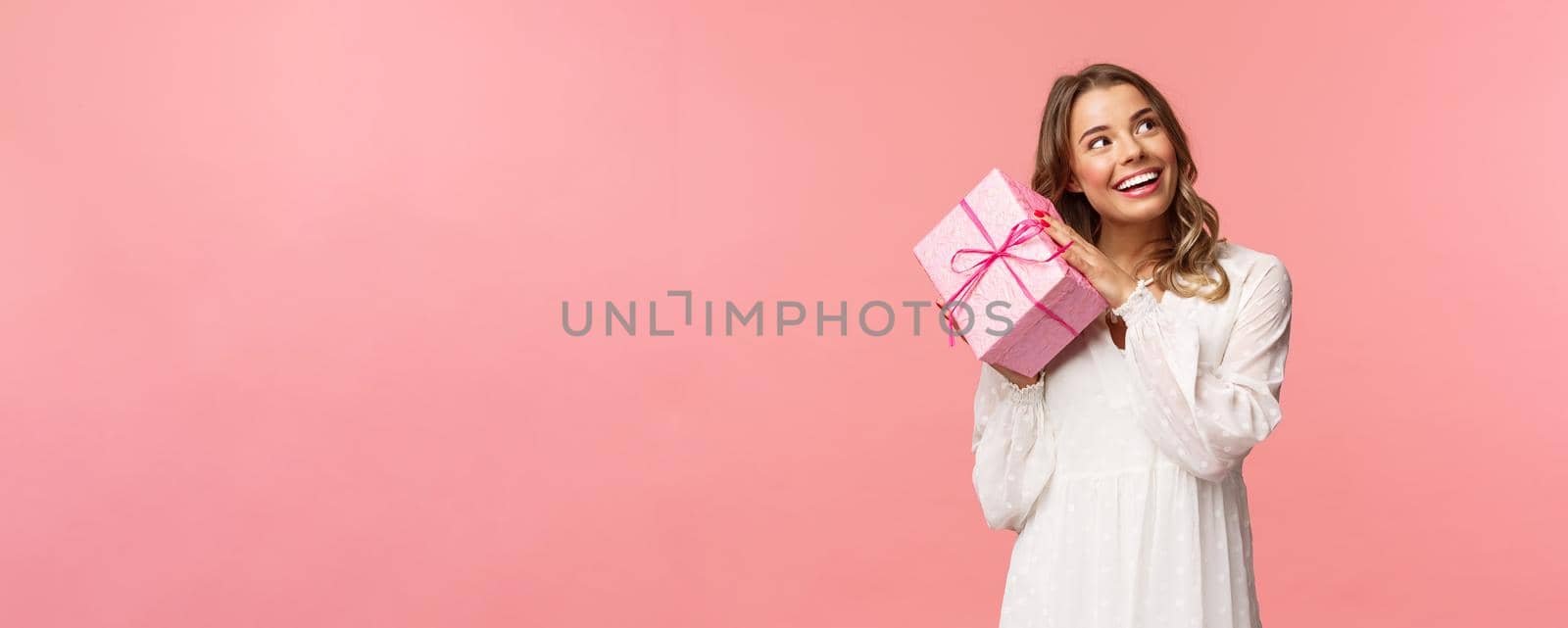 Holidays, celebration and women concept. Portrait of curious attractive young blond girl celebrating birthday, shaking pink gift box to found out whats inside, smiling intrigued look camera.