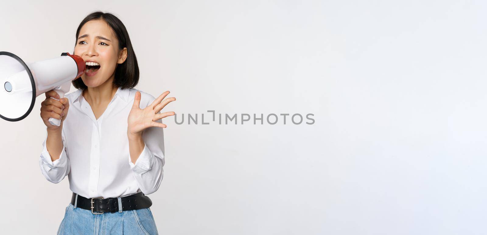 Image of young woman, korean activist, recruiter screaming in megaphone, searching, shouting at loudspeaker, standing over white background.