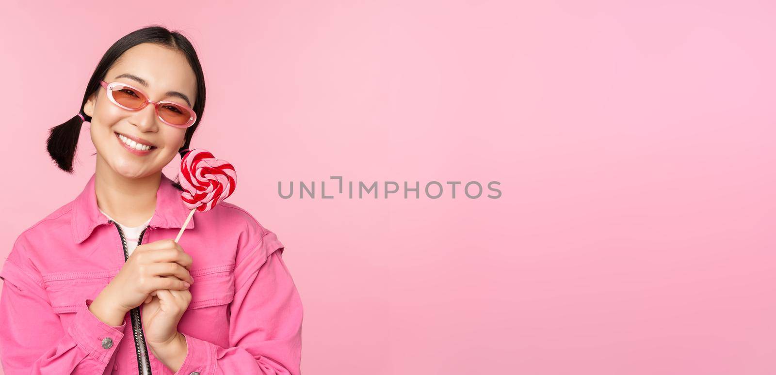 Smiling asian woman in sunglasses, holding lolipop sweets, eating candy and looking happy, standing over pink background.