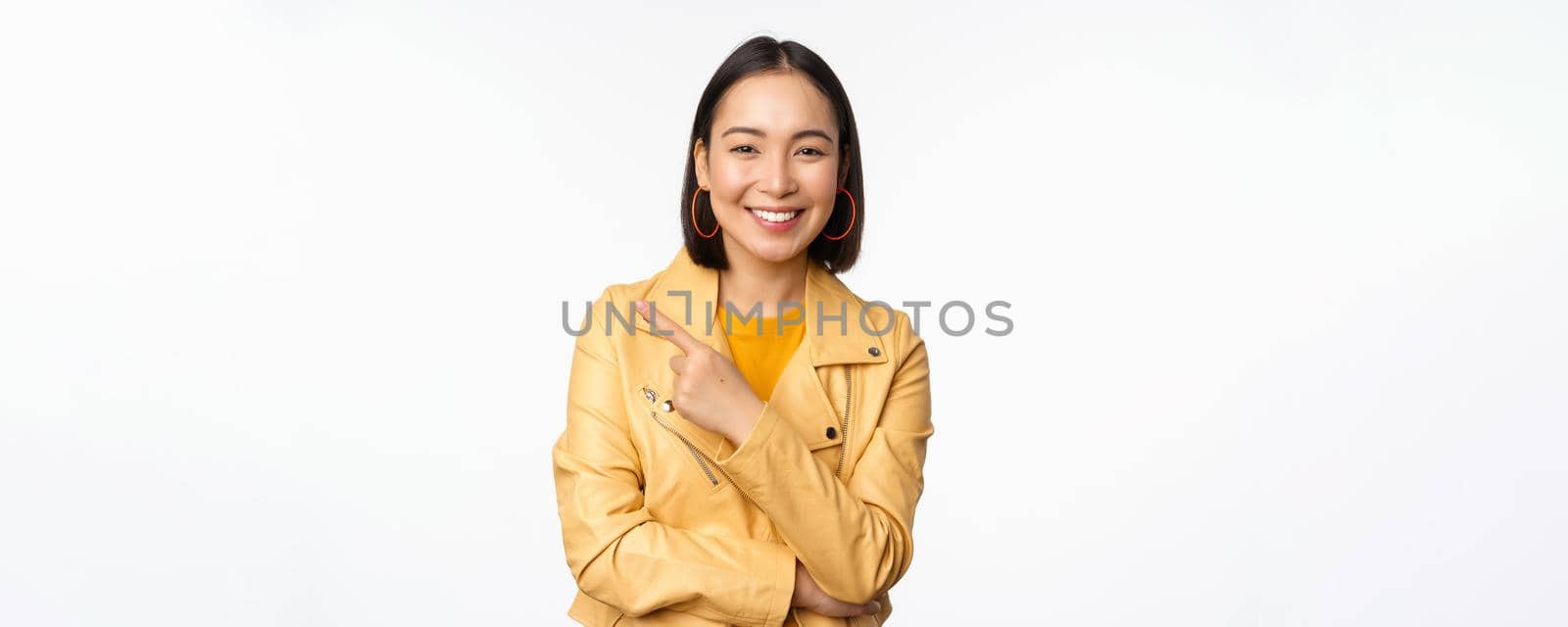 Portrait of smiling asian girl pointing finger left, showing logo or copy space, advertisement on empty side, standing over white background.