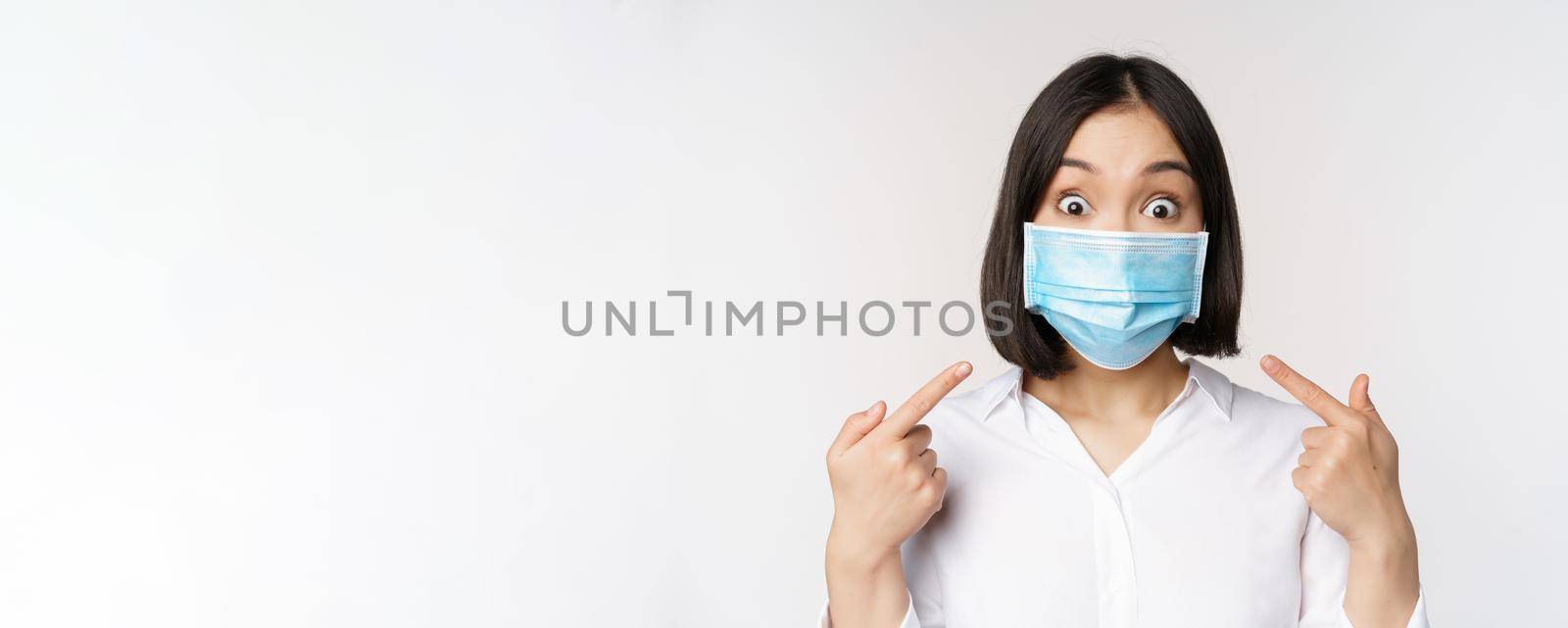 Image of amazed young asian woman pointing at herself while wearing medical face mask, standing over white background.