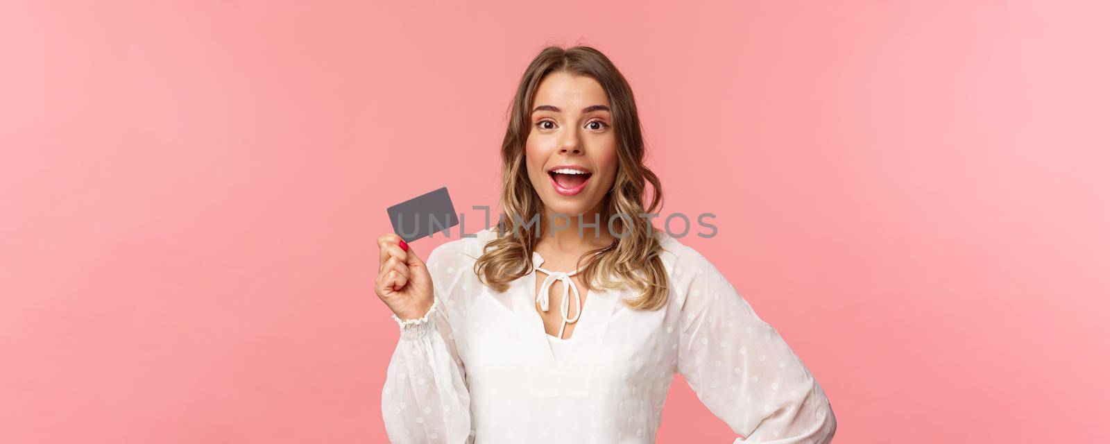 Close-up portrait of excited pretty young blond woman in white dress, holding credit card, showing people how she invests, look camera amused, smiling recommend shopping app, pink background by Benzoix