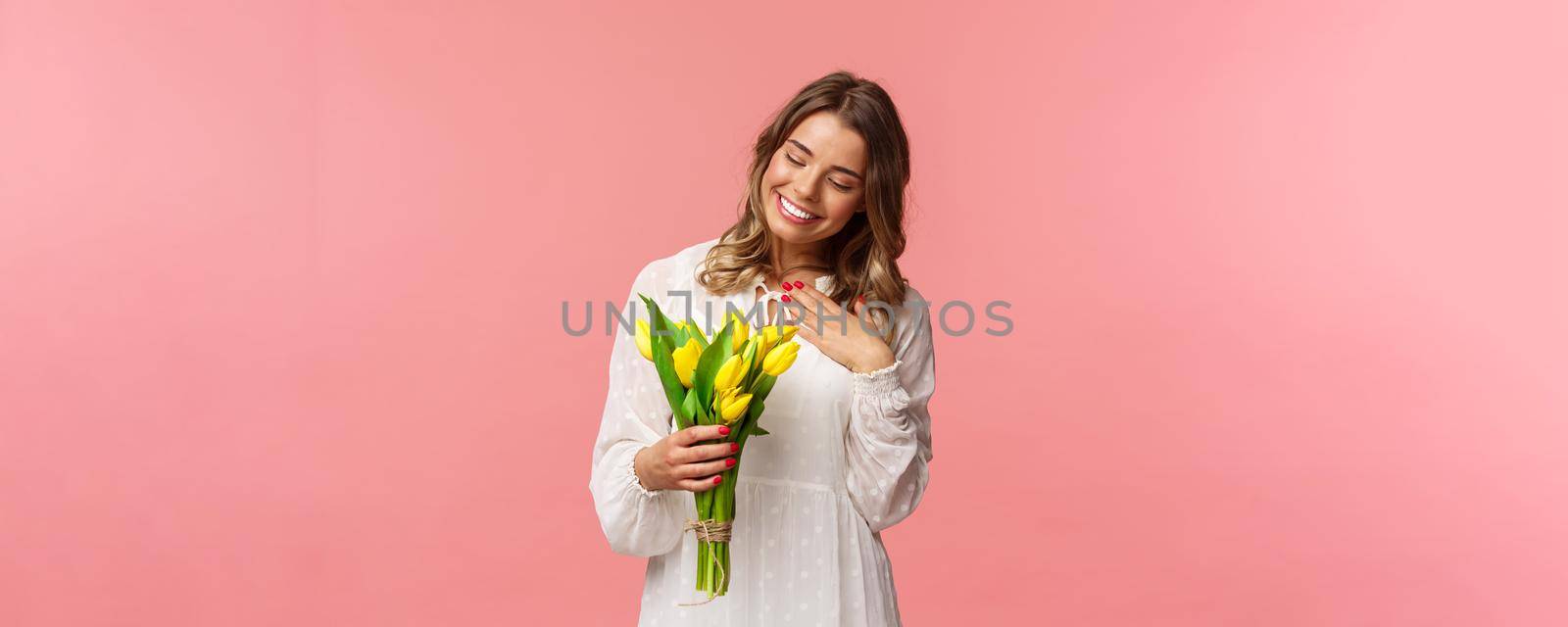 Holidays, beauty and spring concept. Portrait of pleased, cute romantic blond girl receive beautiful bouquet of flowers, looking at yellow tulips and touch heart grateful, smiling.