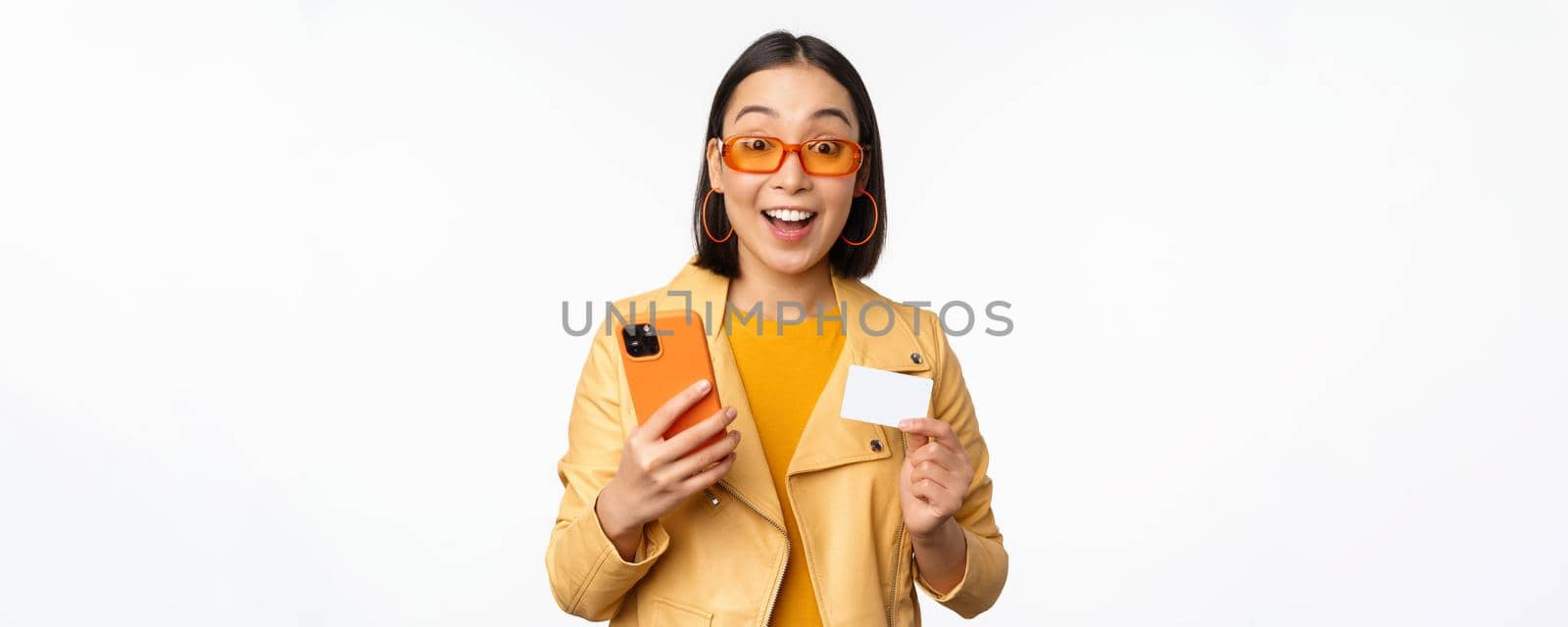 Online shopping. Stylish asian female model in sunglasses, holding credit card and mobile phone, smiling happy, standing over white background. Copy space