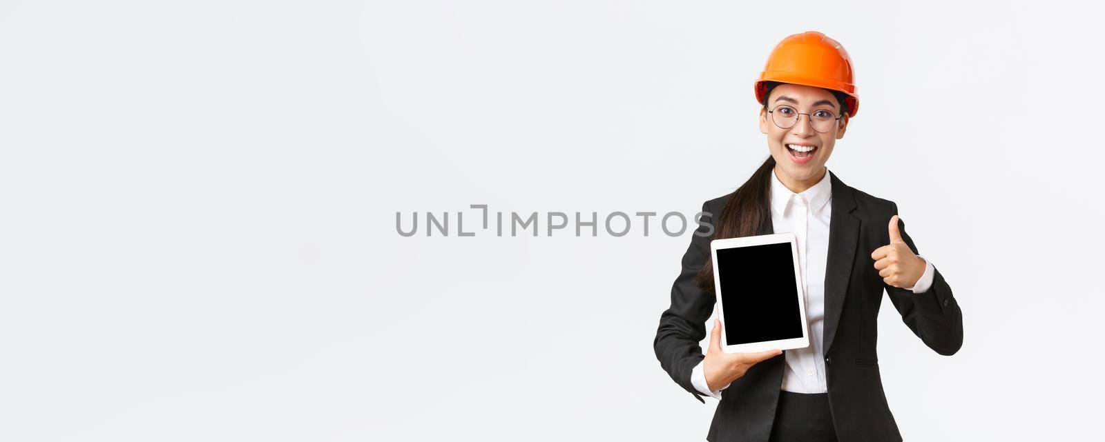 Impressed young female realtor, estate agent showing building during construction, wear safety helmet and business suit, holding digital tablet and make thumbs-up, recommend purchase.