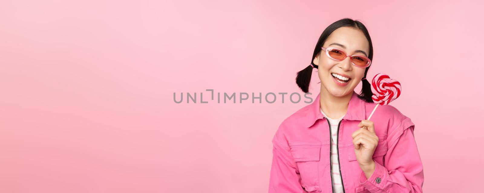 Stylish korean girl licking lolipop, eating candy and smiling, standing in sunglasses against pink background.