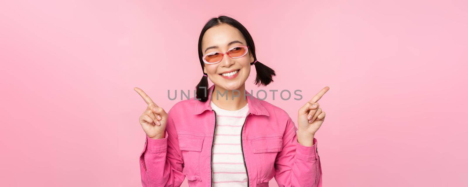 Choice. Stylish korean girl, asian female model points fingers sideways, shows two variants, product advertisement, demonstrating items, standing over pink background.