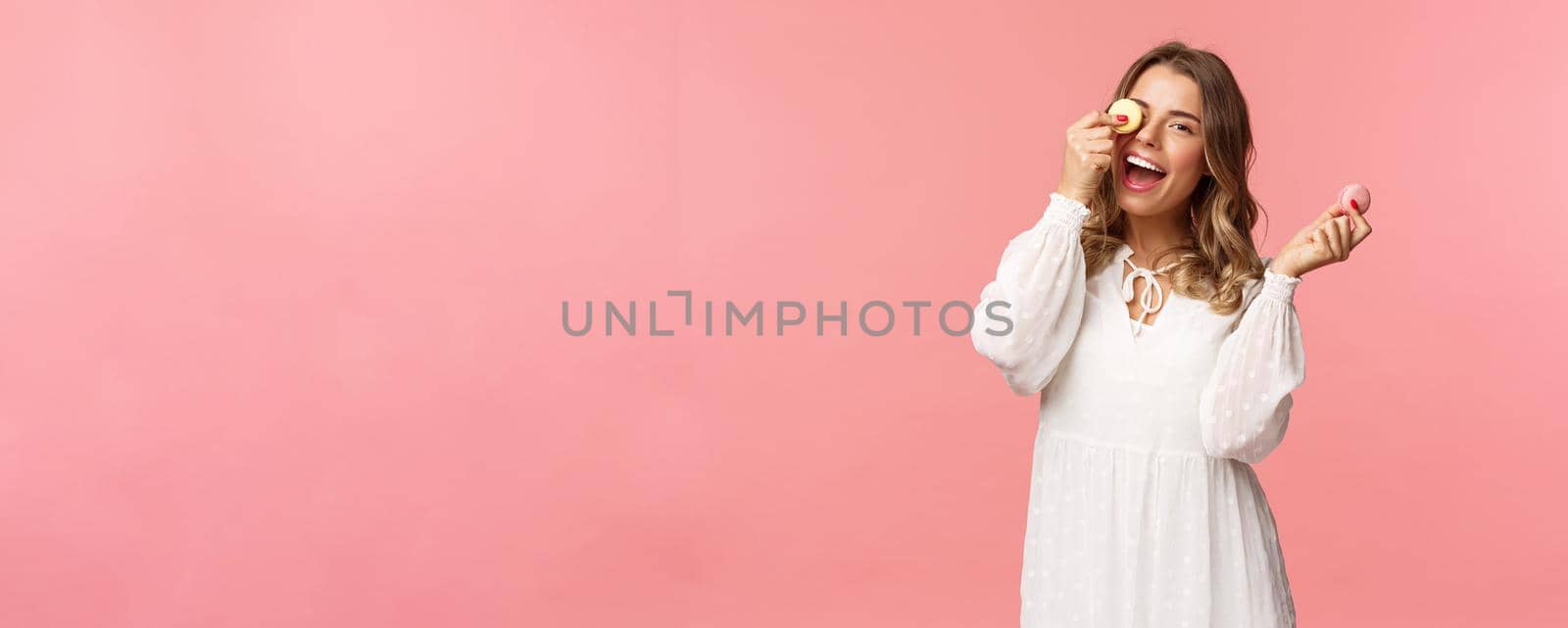 Holidays, spring and party concept. Portrait of carefree, tender and feminine girl in white dress, holding macarons dessert over eye, feeling cheerful, eating sweets, tasty desserts, pink background.