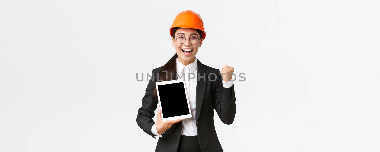 Successful winning female engineer at factory, construction manager in safety helmet and business suit fist pump, triumphing over achievement, showing digital tablet display, graph or diagram.