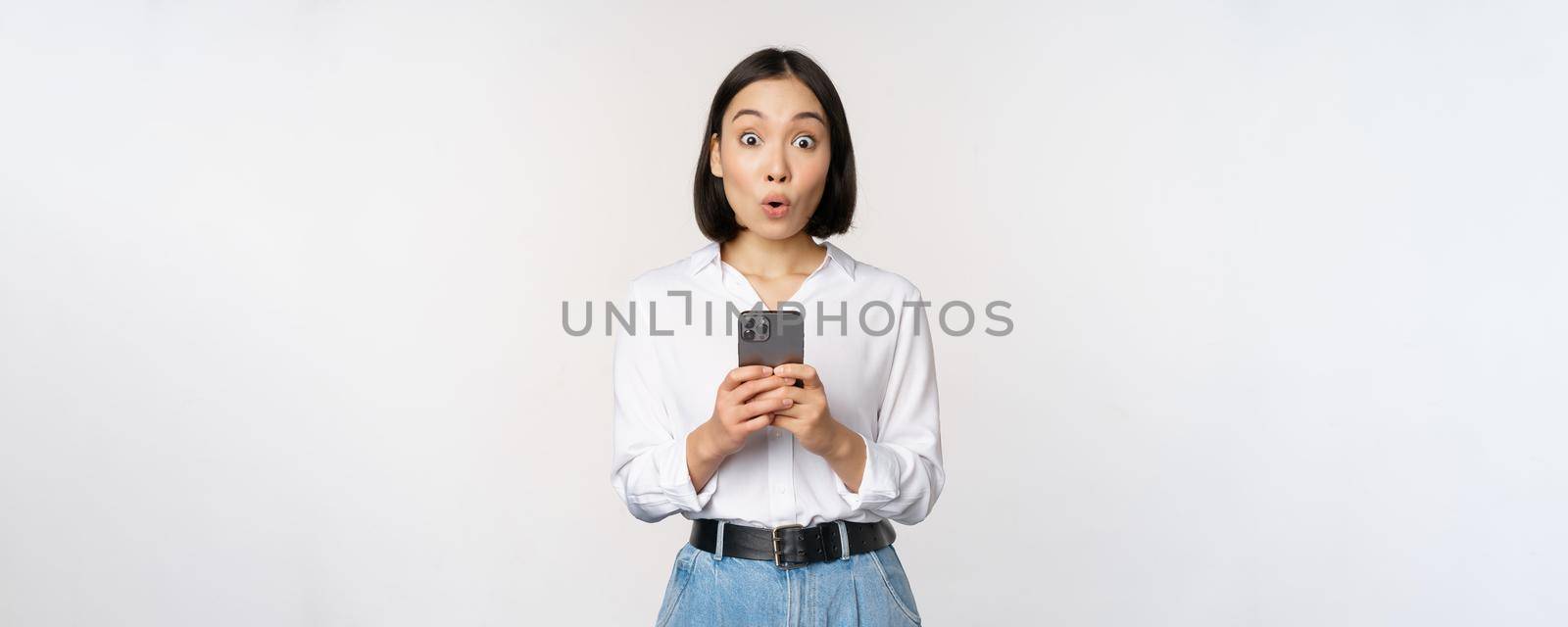 Technology concept. Portrait of asian woman with mobile phone, girl holding smartphone and reacting surprised at cellphone notification, app info, white background.